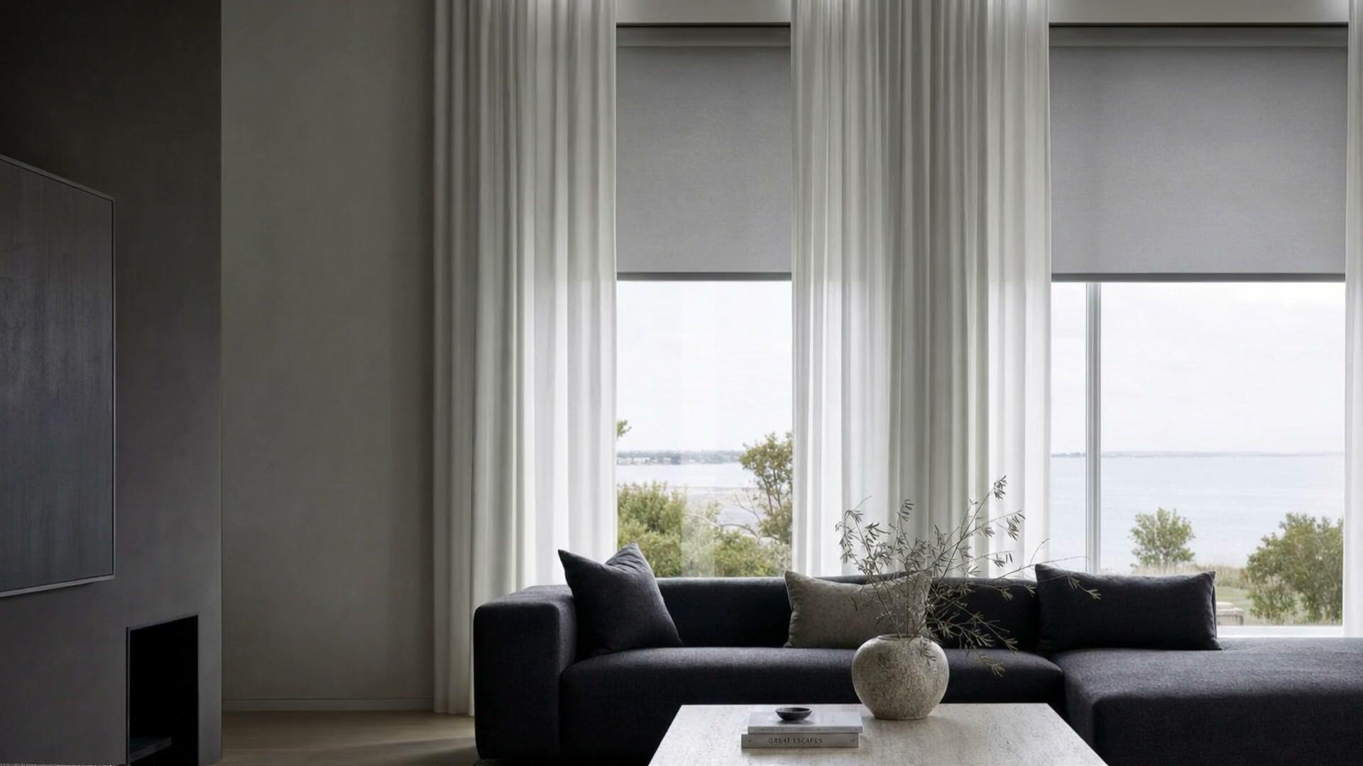 A living room with floor-to-ceiling sheer white drapery and gray roller shades lowered halfway across tall windows, looking out to water