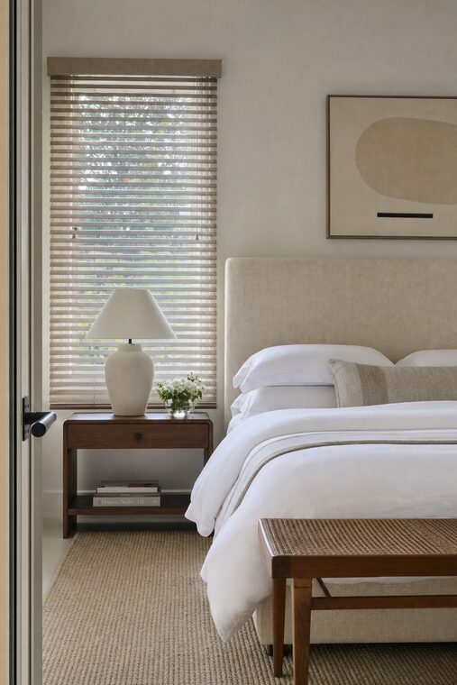A guest bedroom with an oatmeal linen headboard and a caned bench, greige stained wood blinds over the window half closed