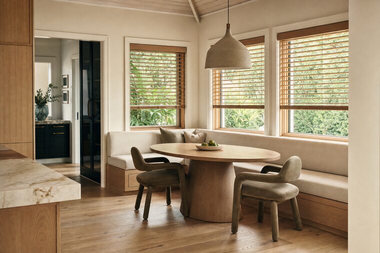 A kitchen breakfast nook with a built-in linen banquette and a round oak table, natural stained wood blinds tilted open across two windows