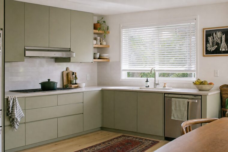 A kitchen with pale green cabinets and open shelving, white faux-wood blinds over the window above the sink with the louvers tilted open