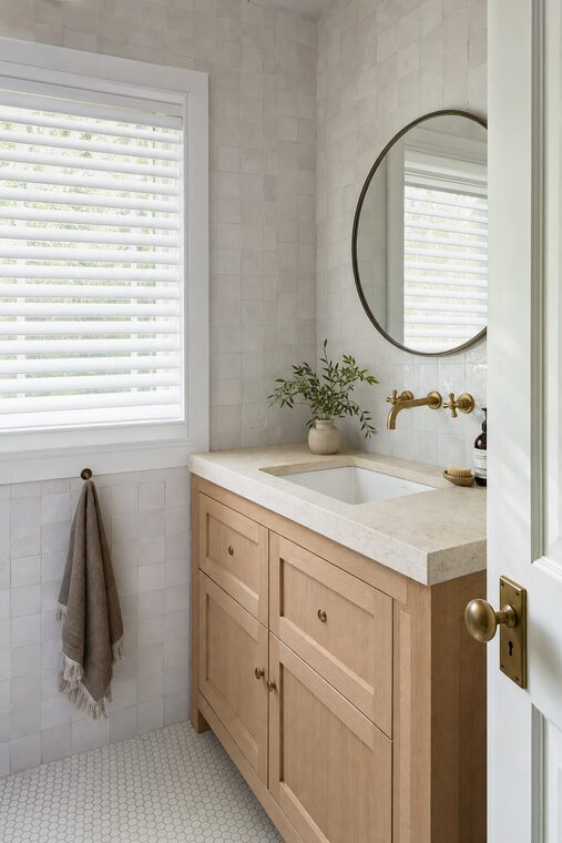 A bathroom with white zellige tile and a pale oak vanity, white painted wood blinds tilted up over the window beside the basin