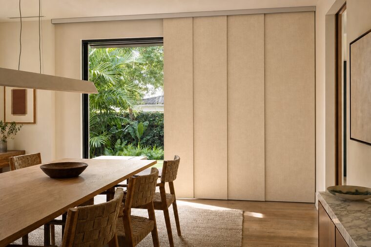 Four wide flax panels on a ceiling track across a dining room slider, one slid back to show the garden