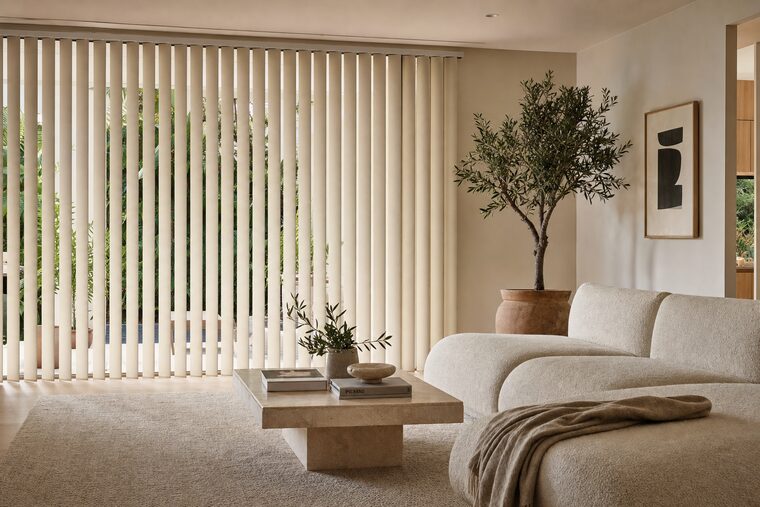 Oatmeal fabric vertical blinds across a wide sliding glass door, vanes tilted so the garden shows in slices between them