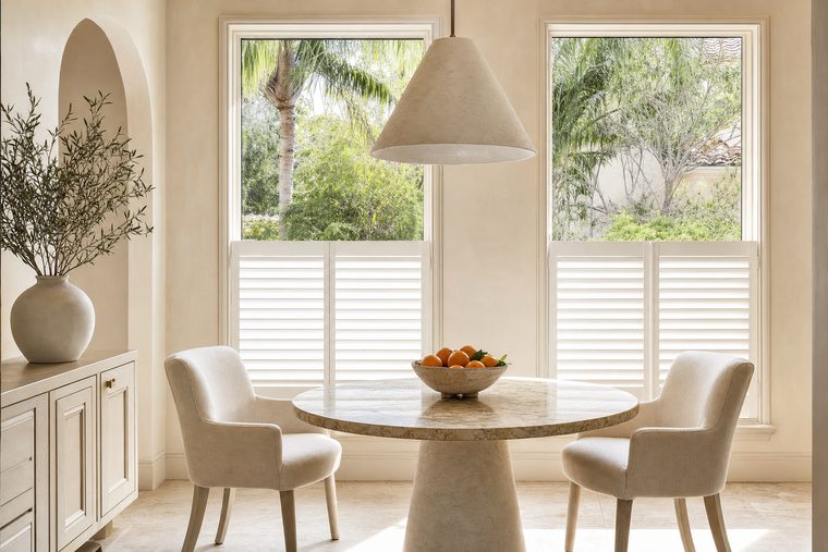 A breakfast room with a round marble table, cafe style shutters covering only the lower half of two tall windows with the tops left open