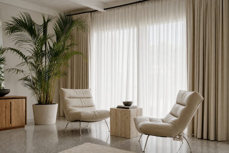A sitting room with cream leather lounge chairs and a potted palm, a sheer white under-curtain drawn across the glass and oatmeal drapery held open either side