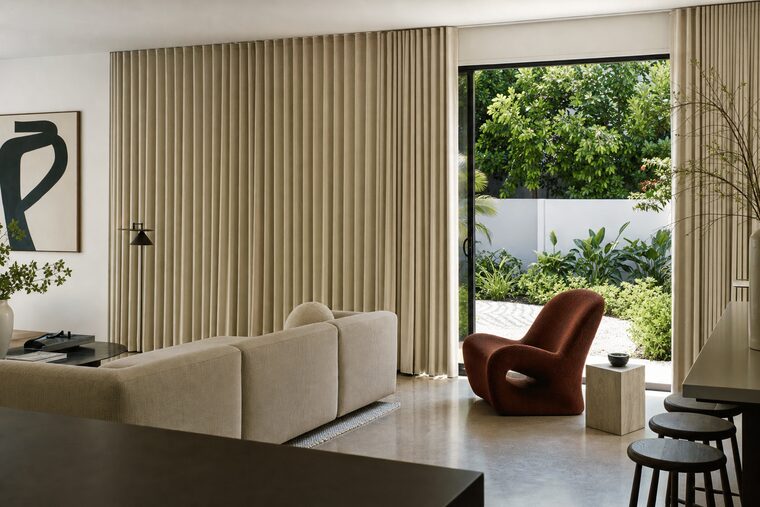 An open-plan living space with a long low sofa and polished concrete floor, oatmeal ripple-fold drapery running wall to wall across sliding doors with one section drawn back