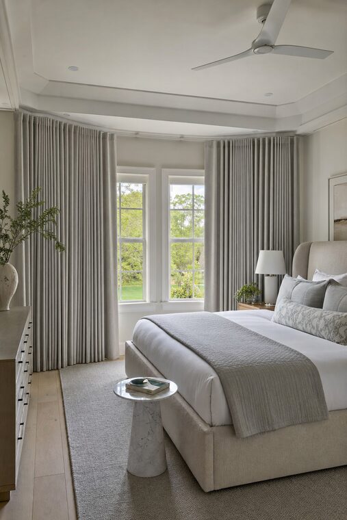 A bedroom with a shallow bay window, an upholstered headboard, and layered stone-colored bedding, soft gray drapery hung across the whole bay to the floor