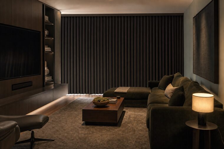 A media room with a dark olive velvet sectional and one lit table lamp, heavy lined charcoal drapery drawn across a wide window