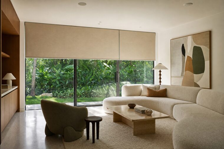 A living room with an ivory boucle sectional and a travertine table, motorized flax roller shades lowered over a sliding door and the window beside it and stopped at the same height
