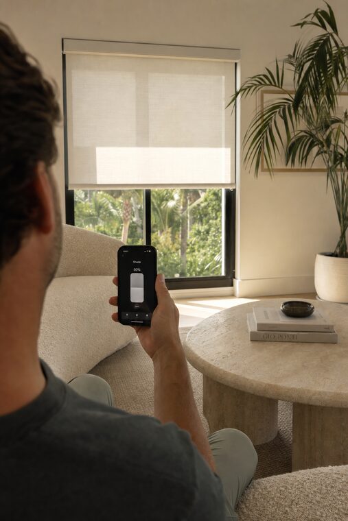 A person holding a phone showing a shade control app, a motorized flax roller shade halfway down a tall window behind them in a living room