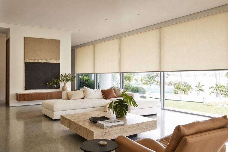 A great room with a low sectional and a travertine plinth table, motorized flax roller shades across a long wall of windows all stopped at the same height