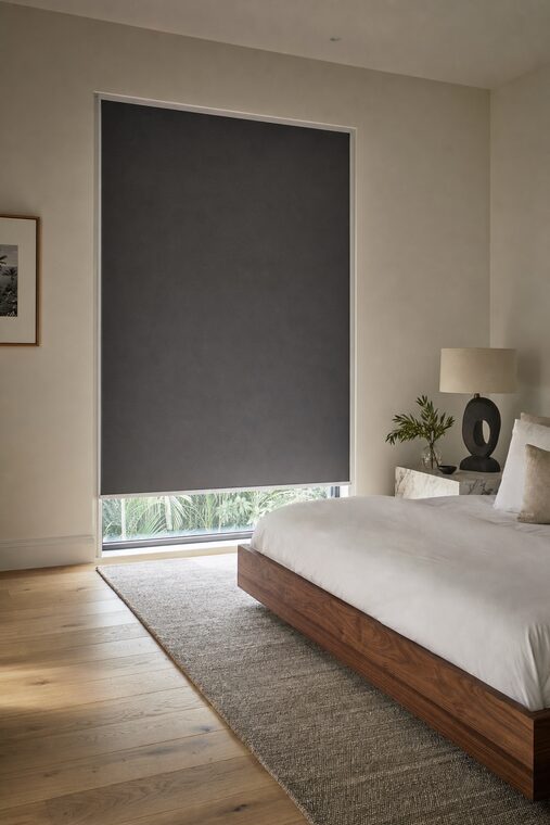 A primary bedroom with a low walnut bed and a marble bedside table, a charcoal motorized blackout shade lowered almost fully with a thin line of daylight beneath the bar