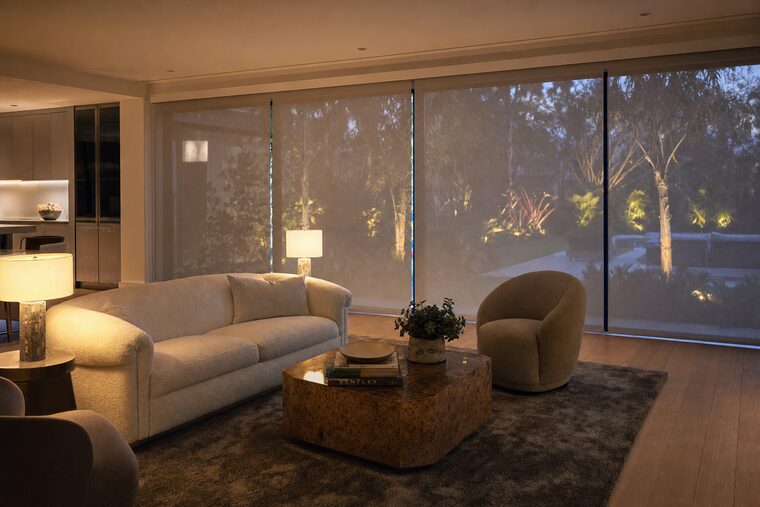 A living room at dusk with a boucle sofa and two lit table lamps, motorized sheer shades lowered across a wall of sliding doors with garden lights glowing through