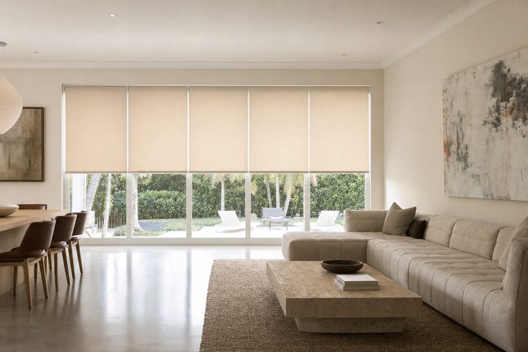 An open-plan great room with a low sectional and a travertine plinth table, five pale flax roller shades across a long wall of windows all stopped at the same height