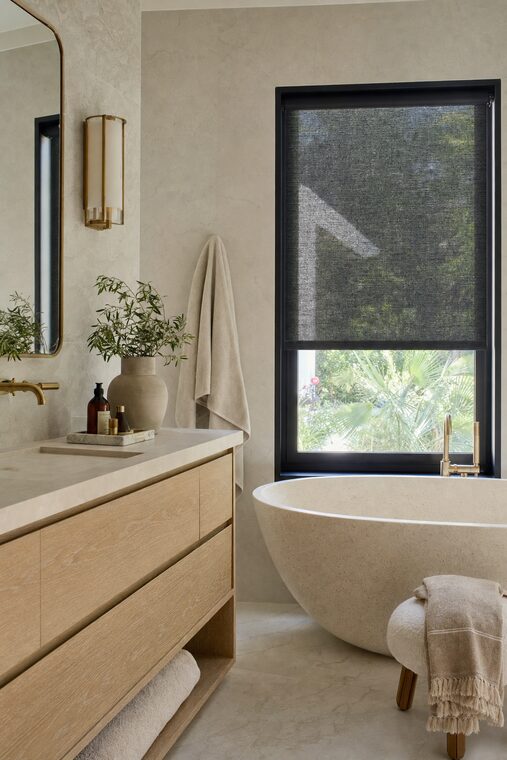 A primary bathroom with a freestanding stone bath and honed marble walls, a charcoal solar shade lowered over the window beside it