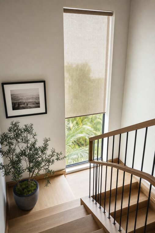 A stair landing with a white oak stair and a blackened steel balustrade, a bone solar shade lowered two thirds over a tall narrow window