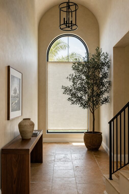A tall entry hall with warm stucco walls and a walnut console, a very high arched window fitted with a bone linen shade cut to the arch and lowered two thirds
