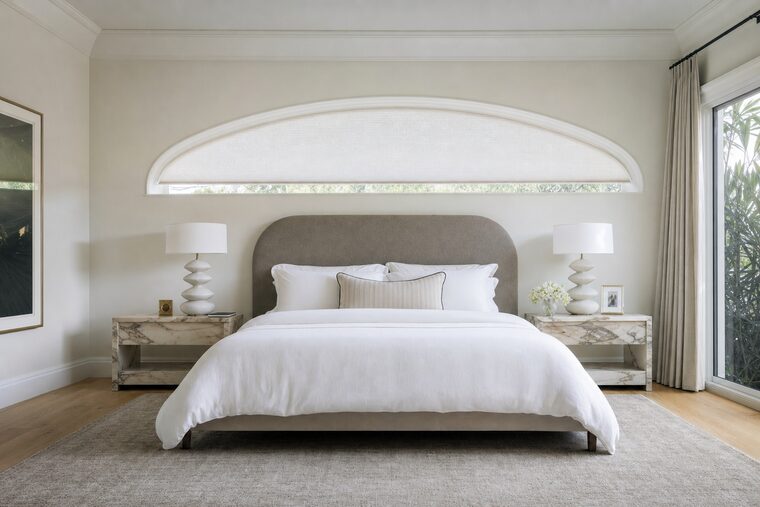 A bedroom with an upholstered headboard and marble bedside tables, a wide low elliptical eyebrow window above the bed fitted with a shade cut to the shallow curve