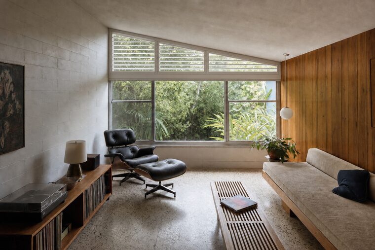 A mid-century ranch living room with a wood-paneled wall and a terrazzo floor, the angled clerestory above the main window fitted with a white plantation shutter cut to the slope of the ceiling