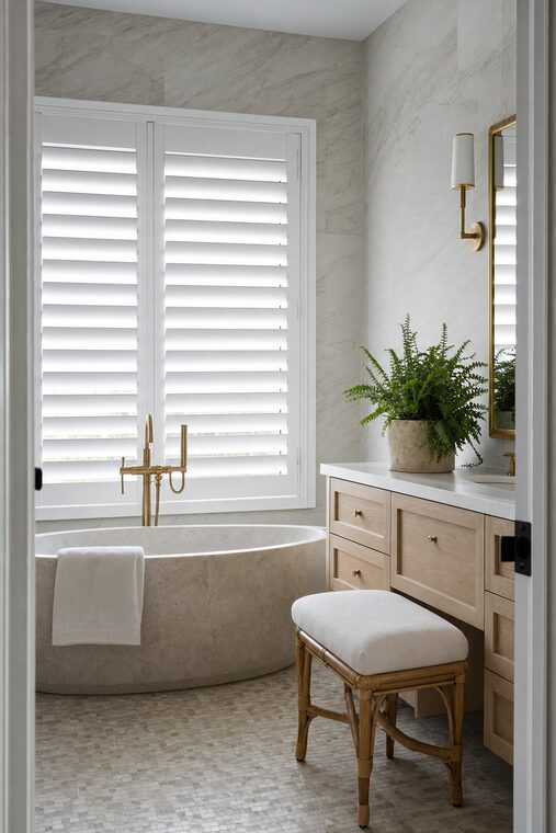 A bathroom with a freestanding stone bath and honed marble walls, white waterproof plantation shutters tilted down over the window beside it