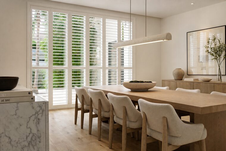 A dining room with a long white oak table and a linear plaster pendant, tall white plantation shutters running floor to ceiling across a run of windows