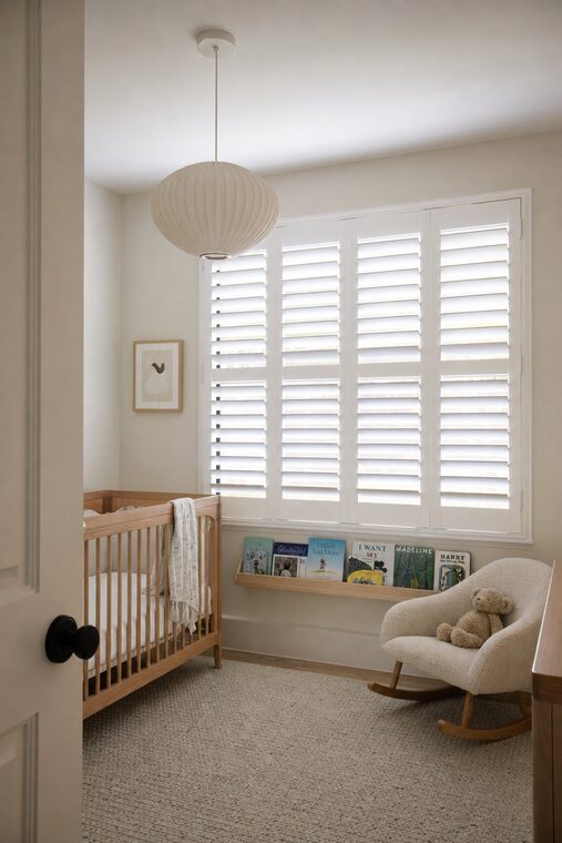 A nursery with a pale oak cot and a boucle rocking chair, white plantation shutters tilted down over the window to soften the light