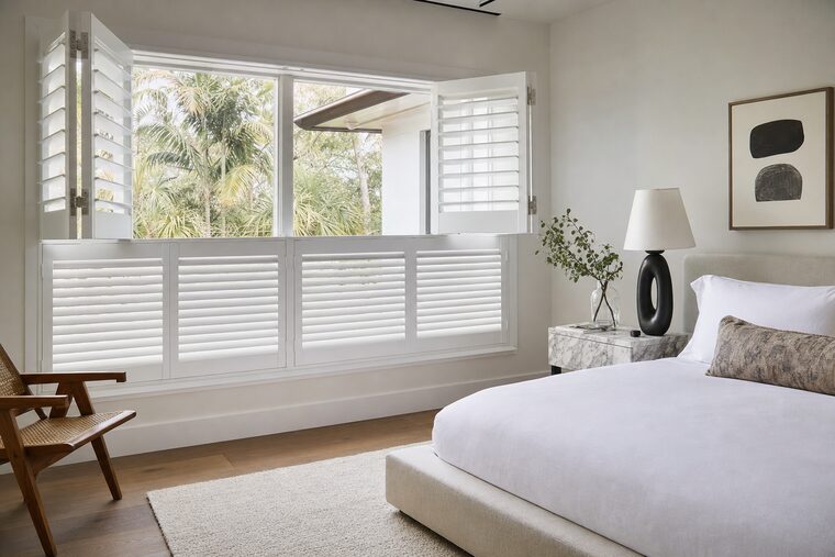 A bedroom with a low upholstered bed, tier-on-tier white shutters across a wide window with the lower tier closed for privacy and the upper tier swung open