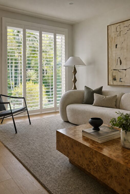 A living room with a boucle sofa and a burl wood coffee table, white plantation shutters across a tall window with the louvers open and level
