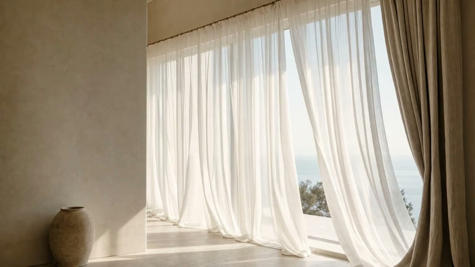 Sheer white drapery drawn across a tall window with a sea view, a linen side panel, and warm afternoon light