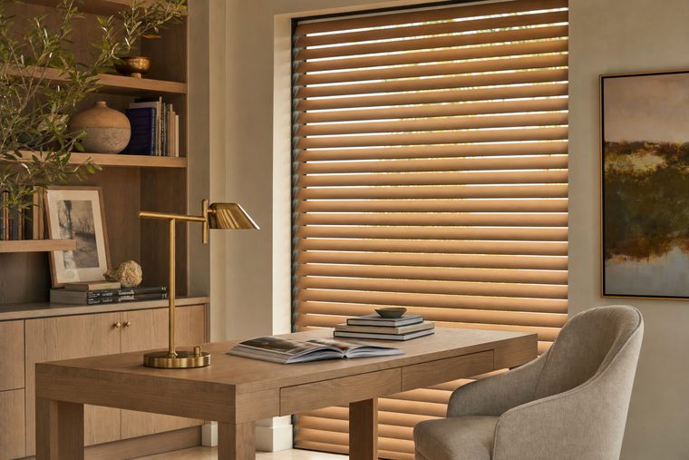 A study in afternoon light with motorized warm oak wood blinds across a tall window behind a white oak desk, louvers tilted down at a precise even angle