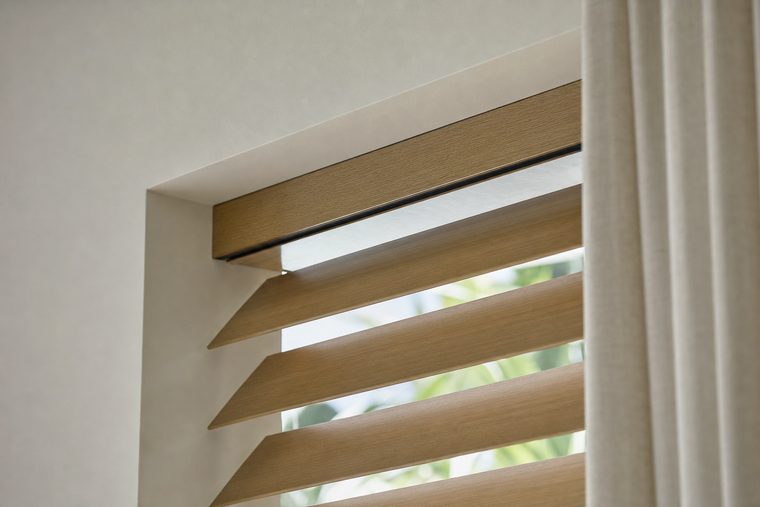 A close detail of the top of a motorized wood blind, a slim oak headrail recessed into a plaster reveal with wide flat oak slats tilted below it