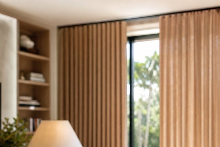 A home office with motorized clay linen drapery half drawn behind a white oak desk and a table lamp