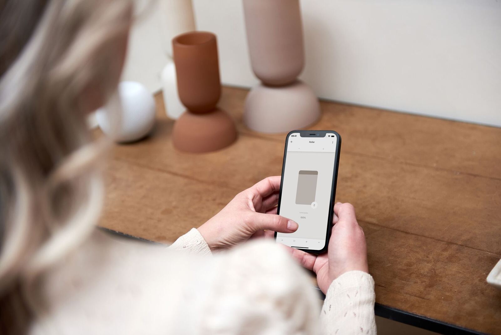 A hand holding a phone with a shade control app open, setting a roller shade's position
