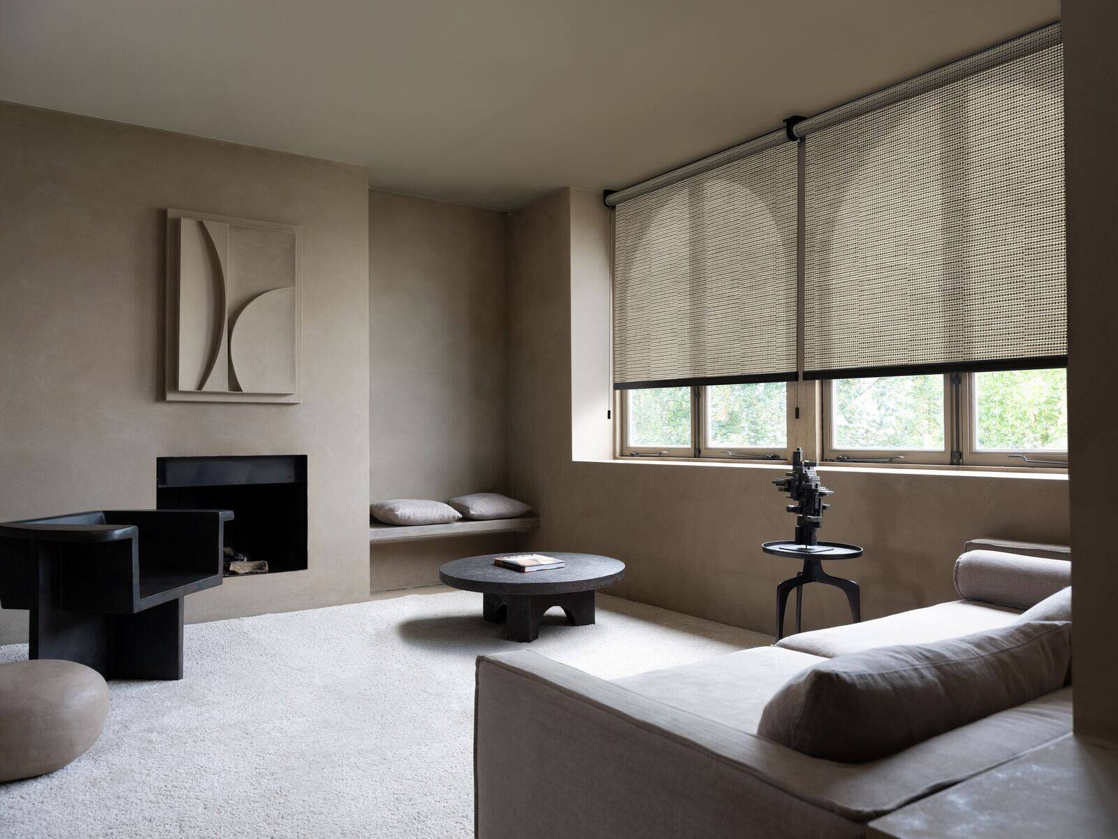 A calm living room in warm stone tones with natural woven roller shades lowered across a run of windows