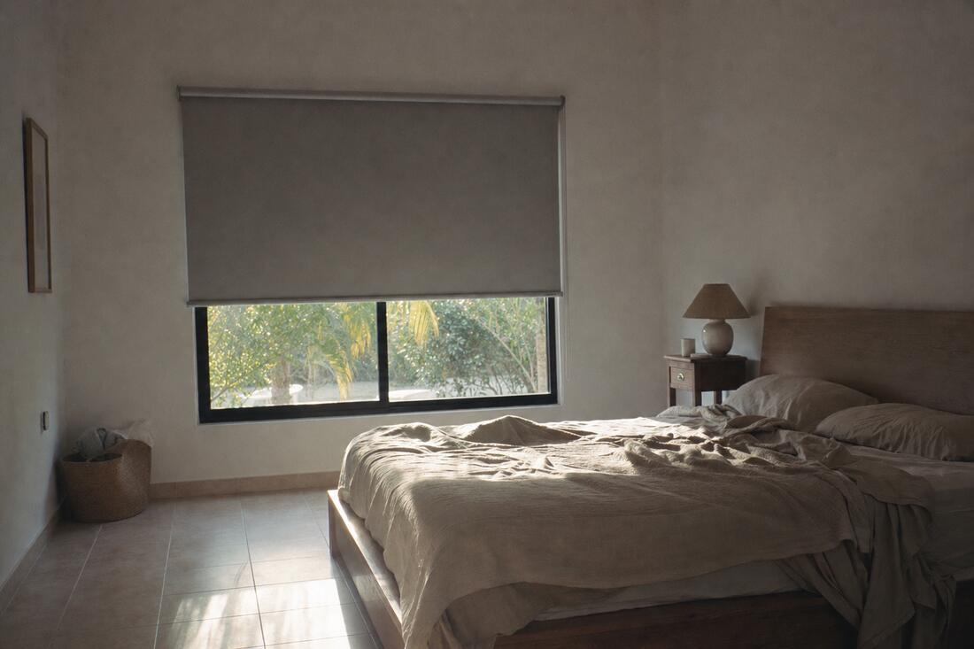 A bedroom at dawn with a gray motorized blackout shade caught a third of the way up, morning light spilling in beneath it