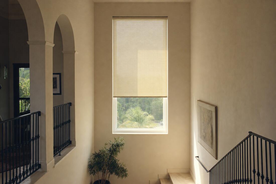 A double-height stair landing with a tall window well out of reach, a bone linen motorized shade lowered two thirds