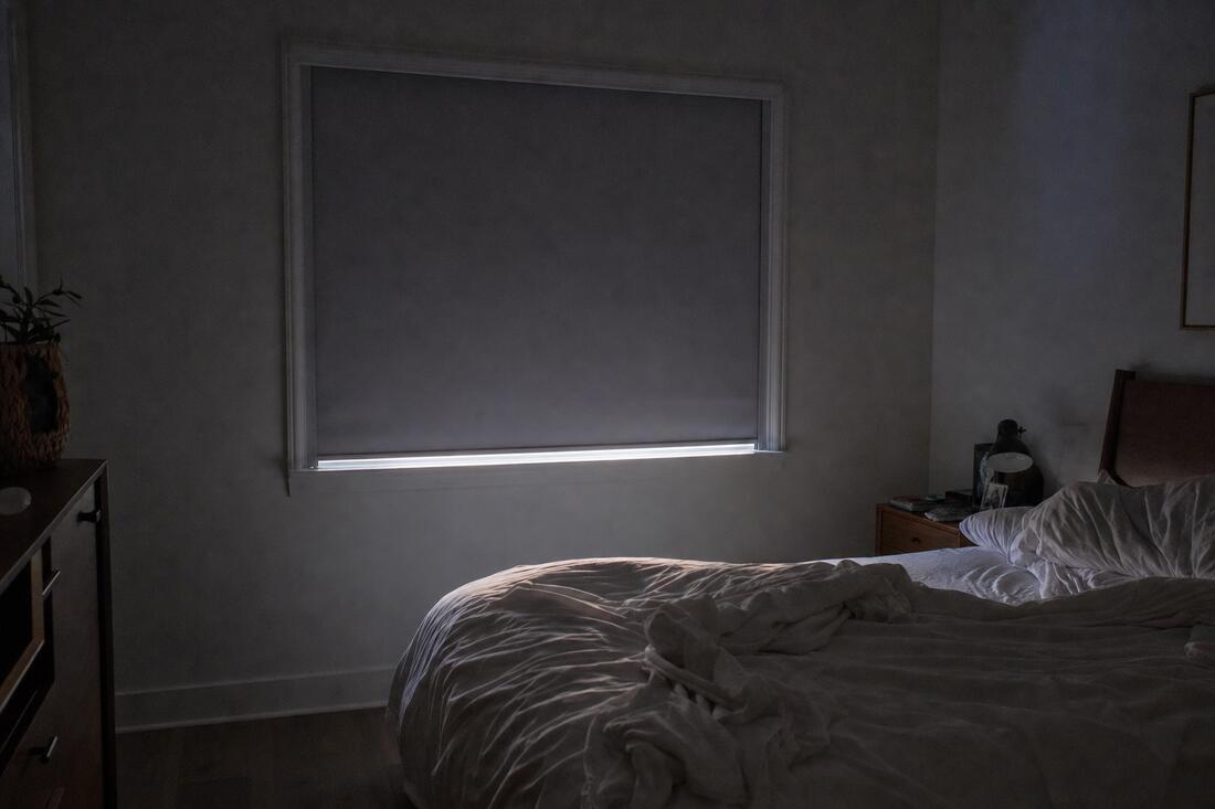 A bedroom in low morning light with a gray blackout roller shade fully lowered in side channels, a thin bright seam along the bottom bar