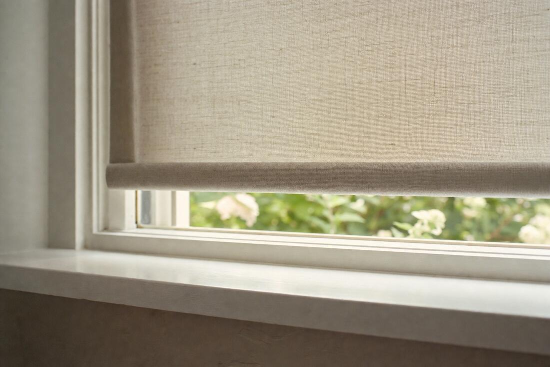 A close view of the fabric-wrapped bottom bar of a roller shade above a white sill, with sunlit garden showing beneath