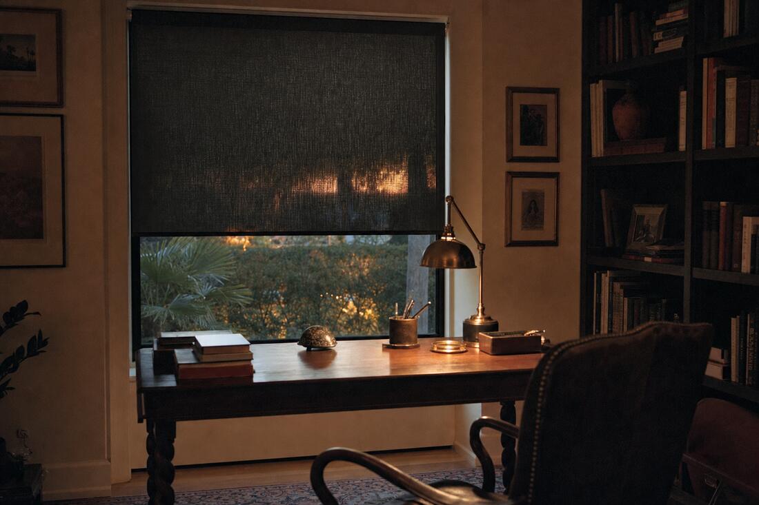 A walnut study at dusk with a lit brass lamp, a charcoal solar-screen roller shade lowered over the window with the garden still visible through the weave