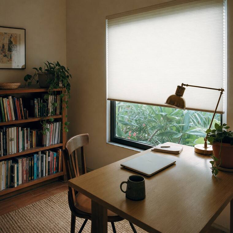 A small home office with a pale oak desk and a brass lamp, a bone cellular honeycomb shade lowered two thirds