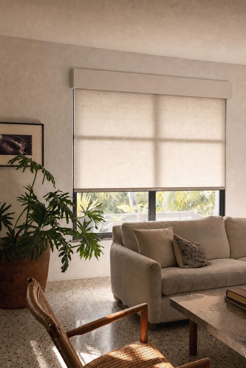 A Florida living room with an ivory sofa and terrazzo floor, an oatmeal linen roller shade lowered two thirds over the window