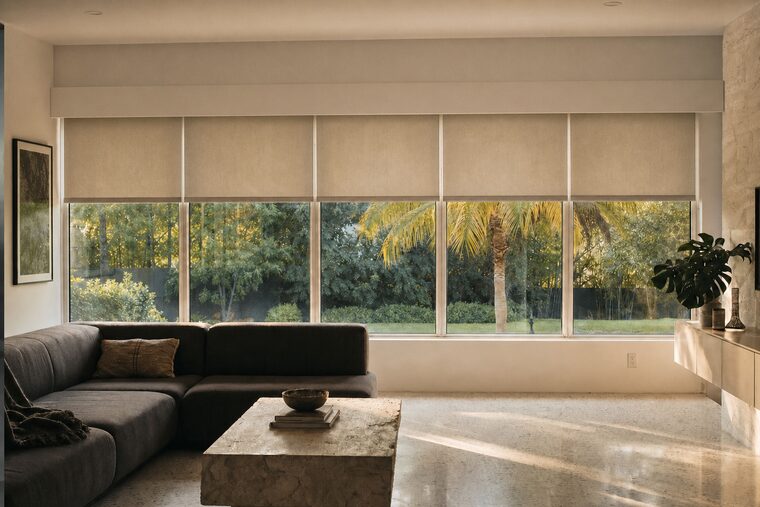 A family room with a long wall of windows, five pale flax roller shades all stopped at the same height in one level line