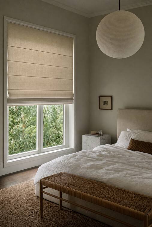 A putty-gray bedroom with white linen bedding and a paper lantern, an ivory roman shade lowered two thirds in flat folds with Florida planting beyond the glass