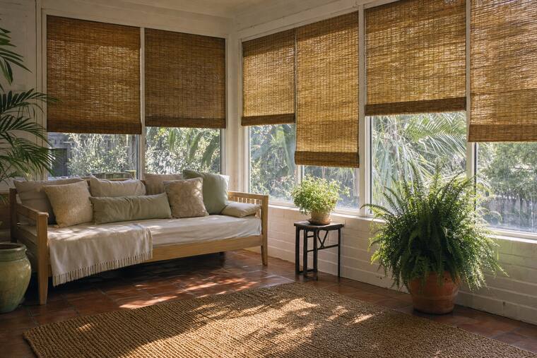 A white-brick sunroom with a daybed and ferns, woven reed shades at varying heights across a run of windows
